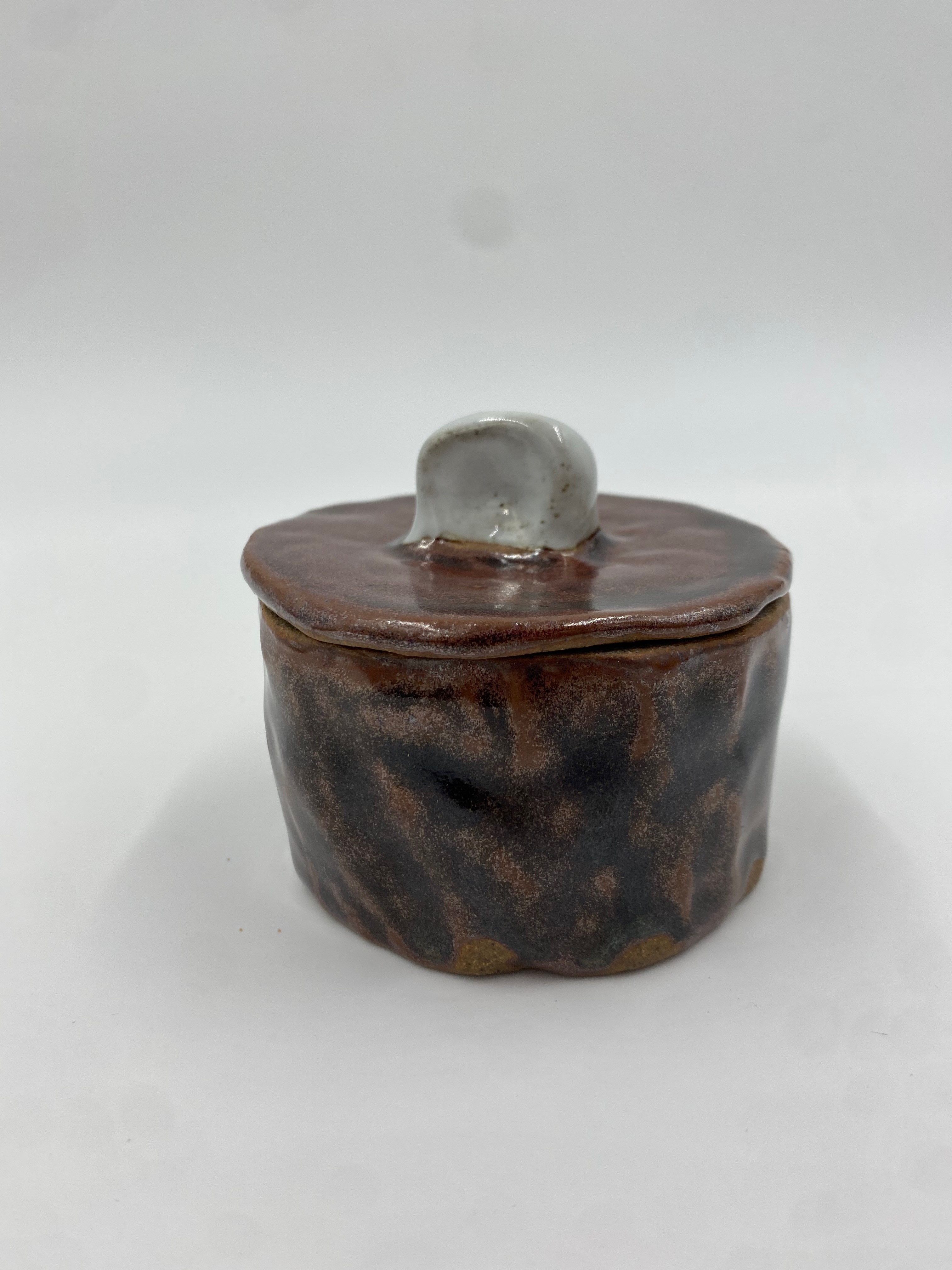 A small pinch-built lidded jar, glazed reddish brown with a white handle on the lid.