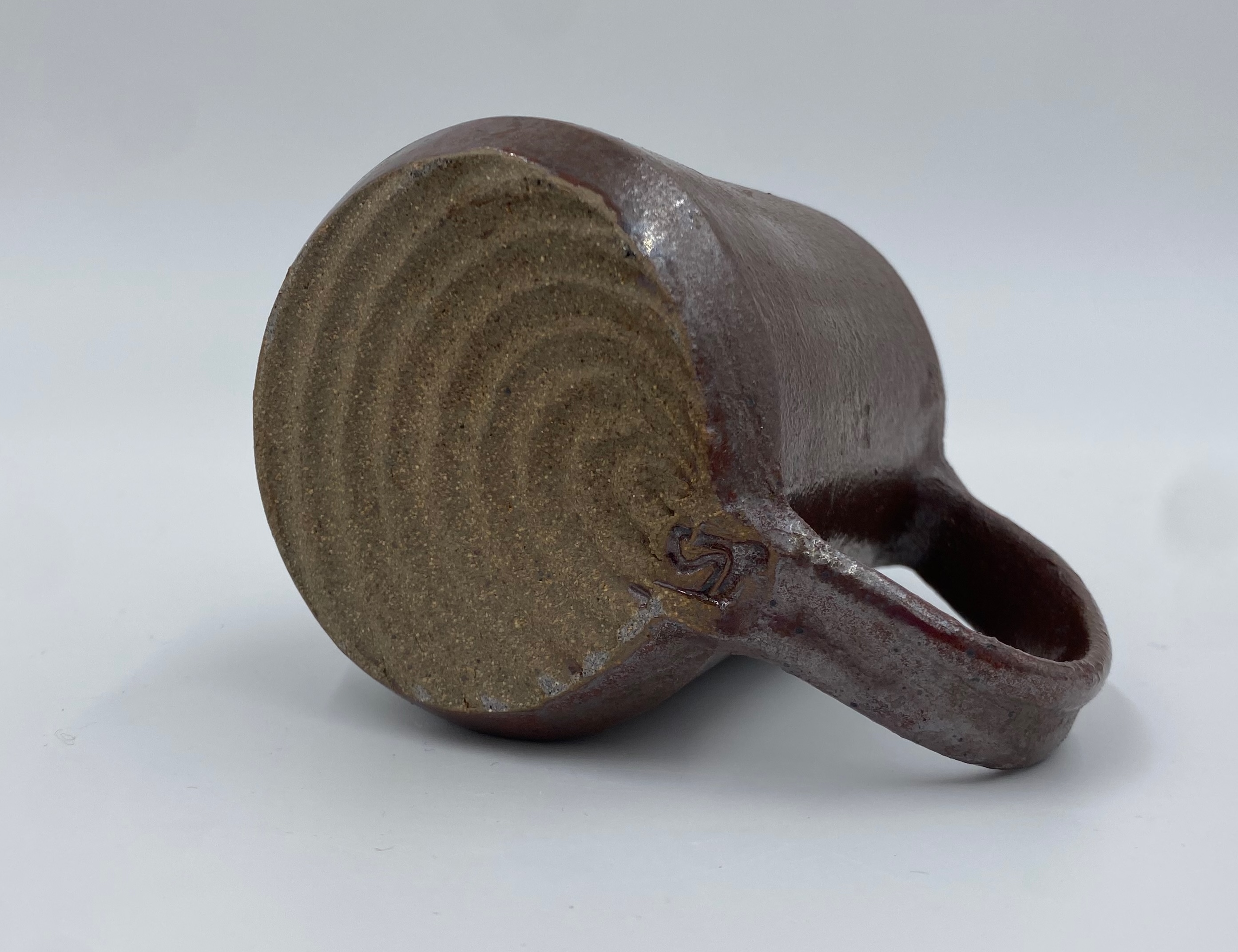 On the base of this metallic red mug is a radial pattern originating at the join-point of the handle. Also at this join-point is a small maker's mark reading r s.