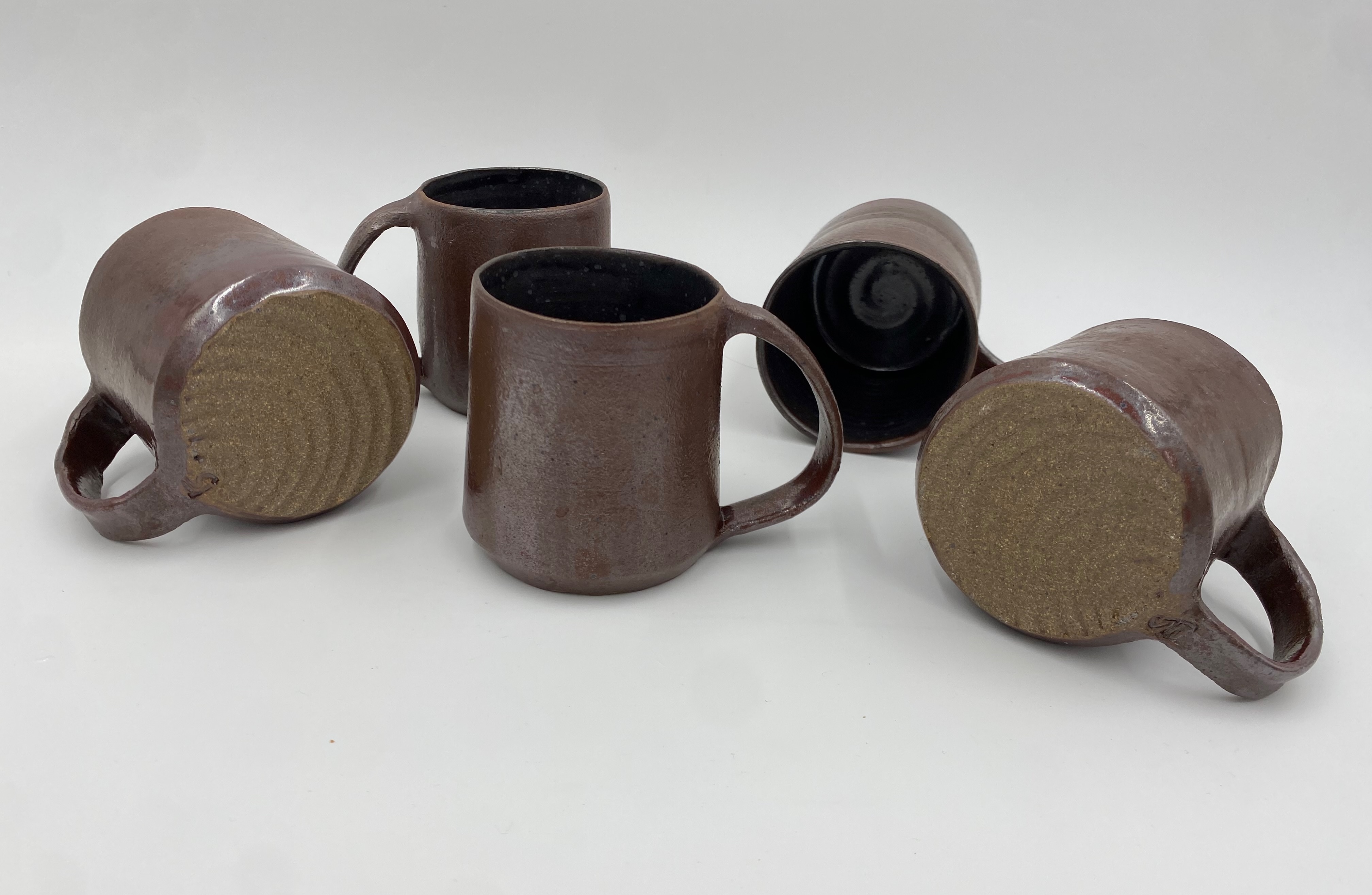 Five metallic red mugs. On one the bottom is visible and has a radial pattern on the base. On another the inside is visible and a subtle spiral is visible through a black glaze.