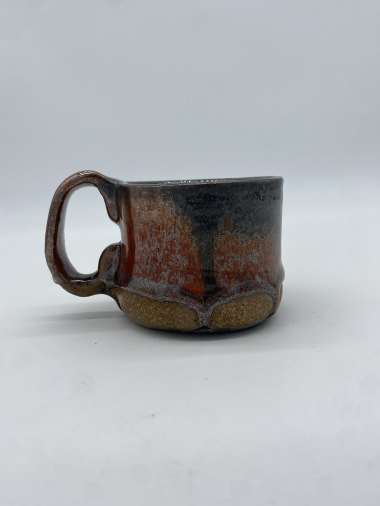 A small stoneware mug with a drippy red-black glaze and a rustic appearance. Some small drips of glaze run to the bottom of the foot.
