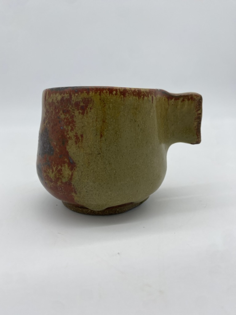 A small stoneware mug with a red-green glaze and a pinched square hand-built handle.